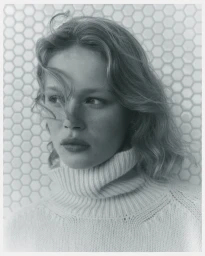Portrait of a female model wearing a cream turtleneck sweater against a patterned tiled background.