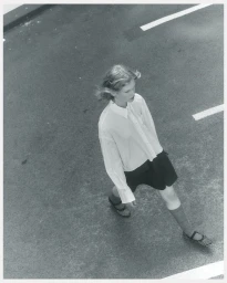 High-angle view of a female model walking across asphalt wearing a white long-sleeve button-down shirt, dark skirt, and flats.