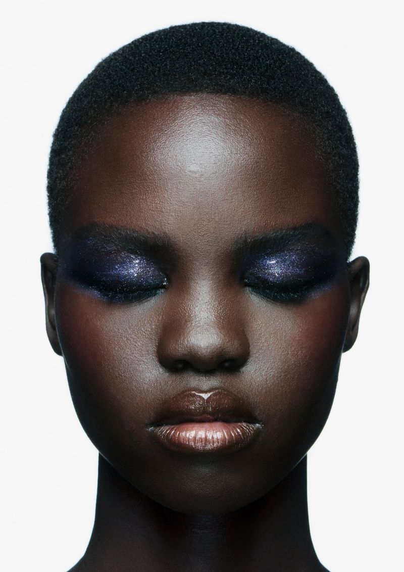 Headshot of a Black woman with dark, glossy skin, closed eyes, and glittery purple eyeshadow against a plain white background.