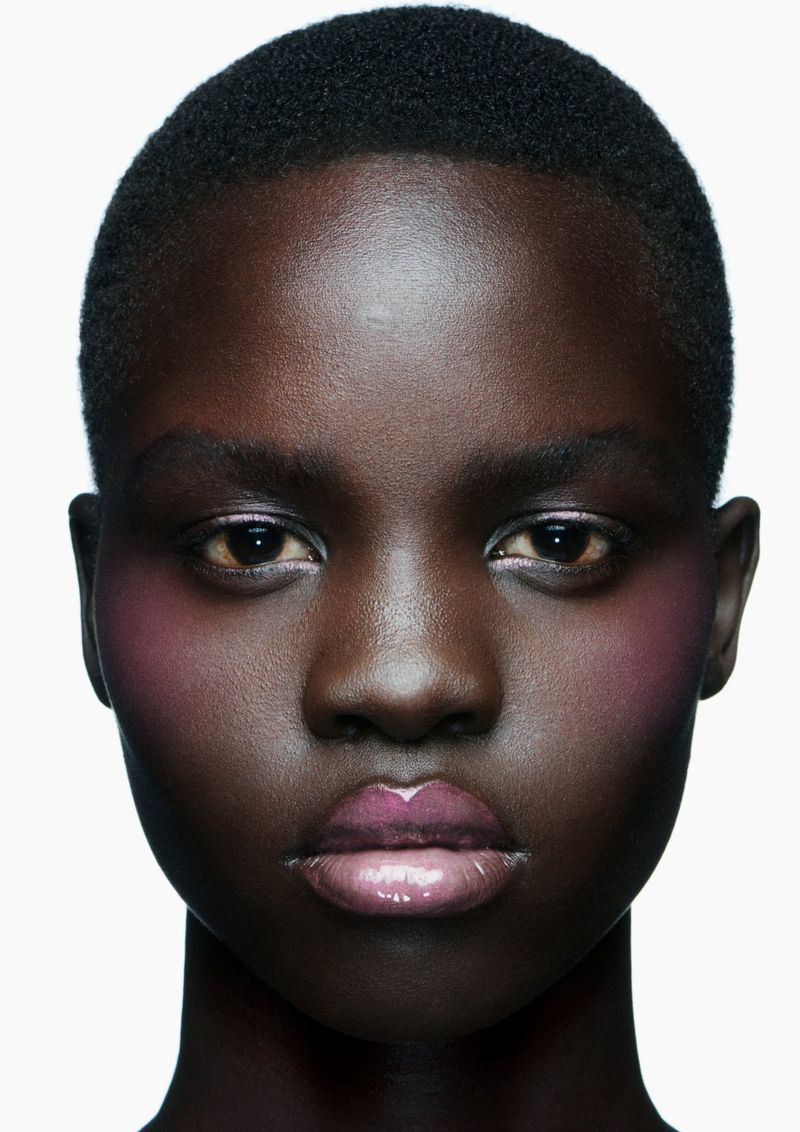 Close-up portrait of a dark-skinned woman with makeup. Light pink blush and glossy lipstick are visible against a plain white backdrop.