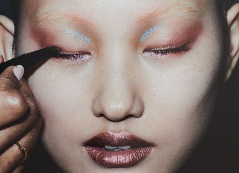 Close-up beauty shot features a model with closed eyes and creative makeup, including copper lipstick, red and silver eyeshadow, and a makeup artist.