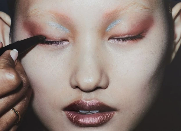 Close-up beauty shot features a model with closed eyes and creative makeup, including copper lipstick, red and silver eyeshadow, and a makeup artist.