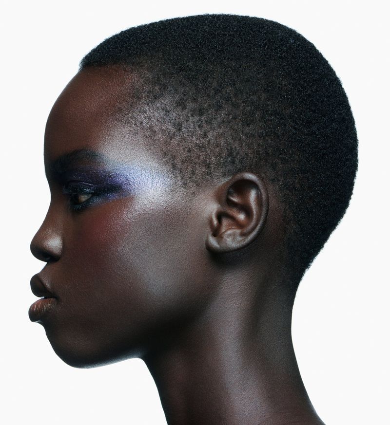 Profile portrait of a dark-skinned adult female with buzz cut hair and eye shadow against a white background in a beauty studio.