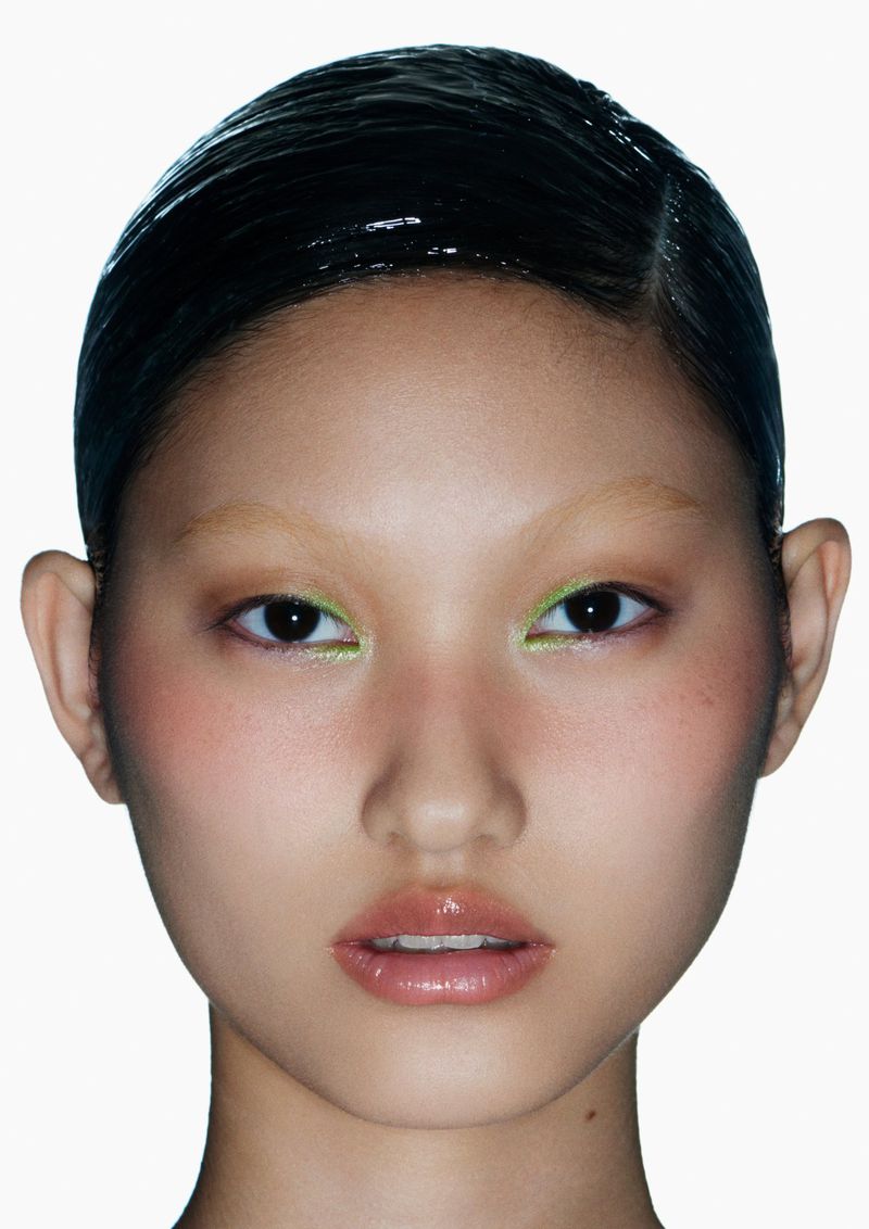 Close-up portrait of an Asian woman with shiny black hair, wearing bright green eyeliner, facing forward against a plain white background.