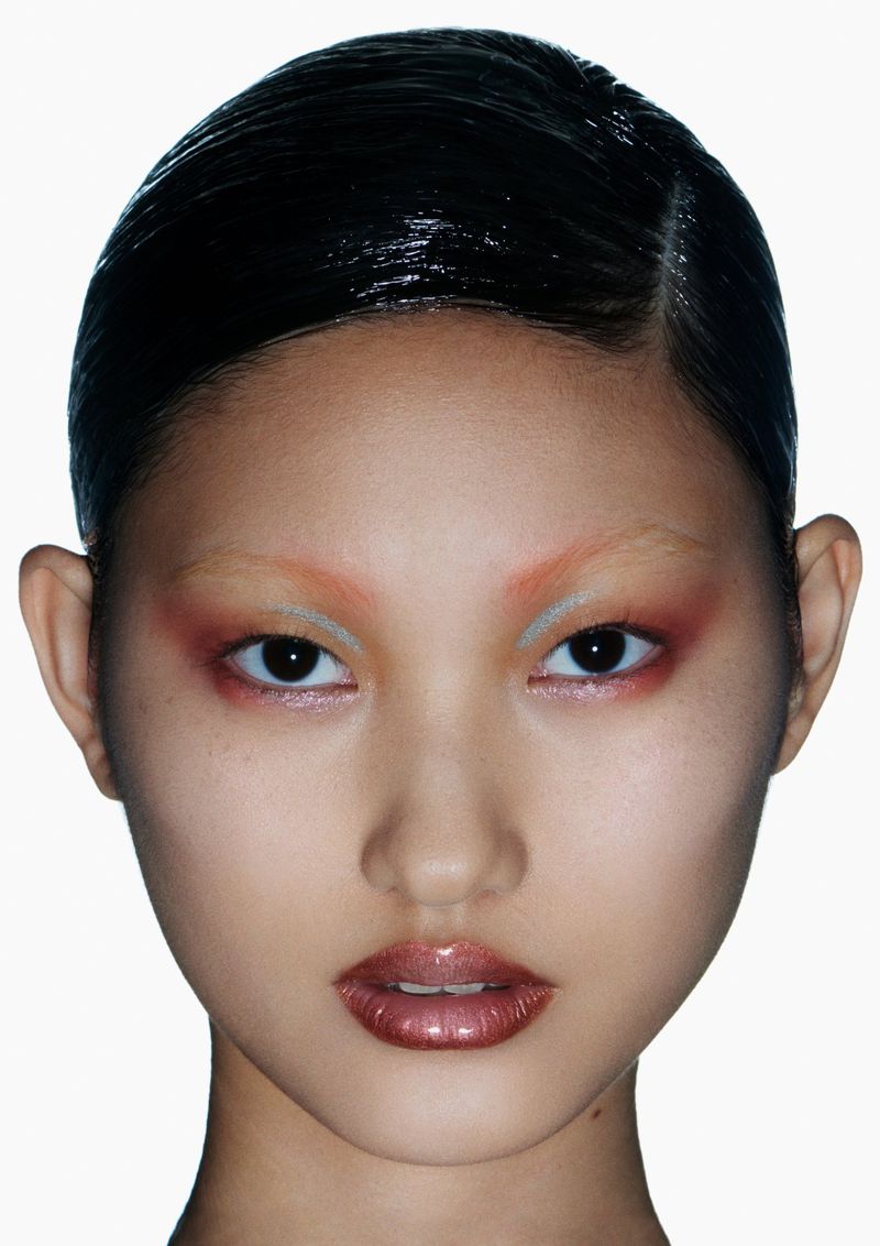 Close-up portrait of a young Asian woman with dramatic coral and silver eyeshadow, glossy lips, and sleek wet-look hair against a white background.