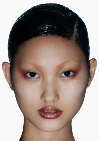 Close-up portrait of a young Asian woman with dramatic coral and silver eyeshadow, glossy lips, and sleek wet-look hair against a white background.