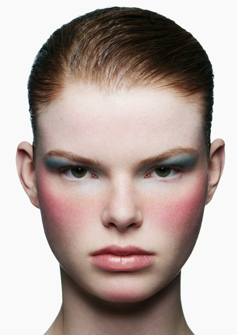 Close-up studio portrait of a redhead adult female with green eyes wearing blue eye shadow and pink blush on a white background.