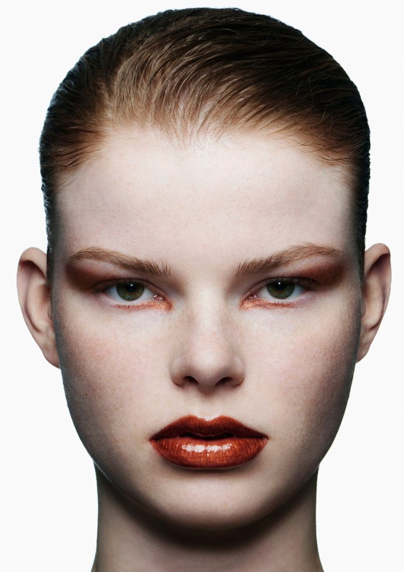 Beauty shot of a woman with slicked-back brown hair, green eyes, and metallic orange makeup on her eyelids and glossy lips in studio lighting.