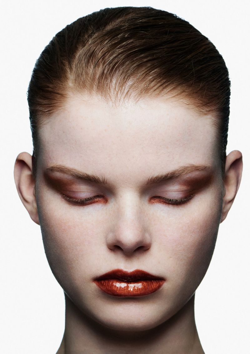 Close-up portrait of a fair-skinned woman with slicked-back brown hair and copper makeup on her eyes and glossy lips against a white background.