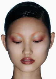 Close-up portrait of a model with slicked-back dark hair, glossy metallic eye shadow, and tinted lips against a white background.