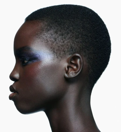 Profile portrait of a Black woman with a shaved buzzcut and shimmering iridescent blue eyeshadow on a white studio background.