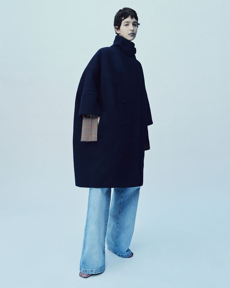 An adult female wearing a dark blue, oversized coat and wide-leg jeans poses in a studio setting, standing against a light blue backdrop.