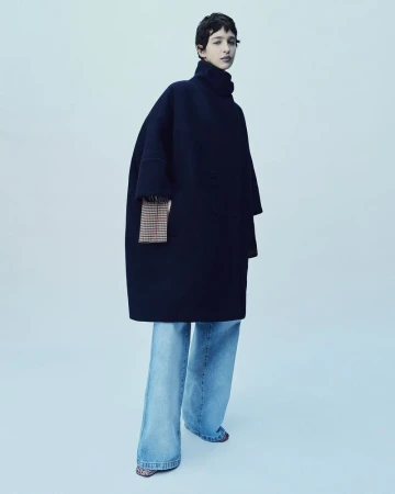 An adult female wearing a dark blue, oversized coat and wide-leg jeans poses in a studio setting, standing against a light blue backdrop.