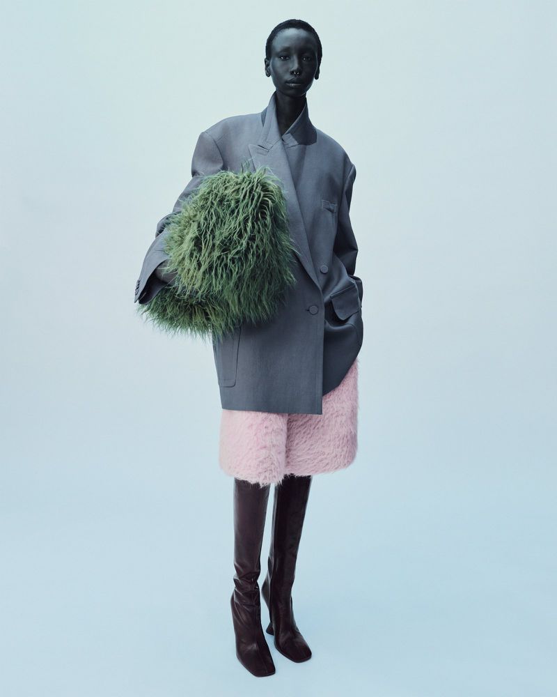 A black woman in an oversized grey blazer, pink shorts, and brown boots poses with a green, furry bag in a studio setting.