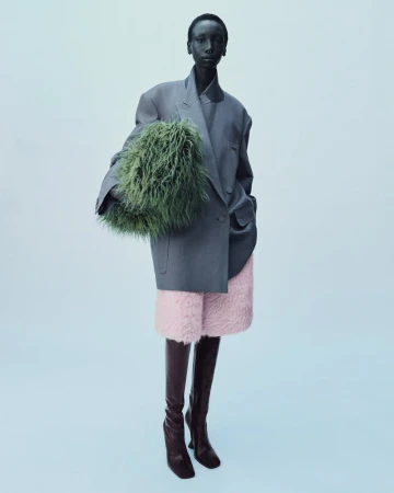 A black woman in an oversized grey blazer, pink shorts, and brown boots poses with a green, furry bag in a studio setting.
