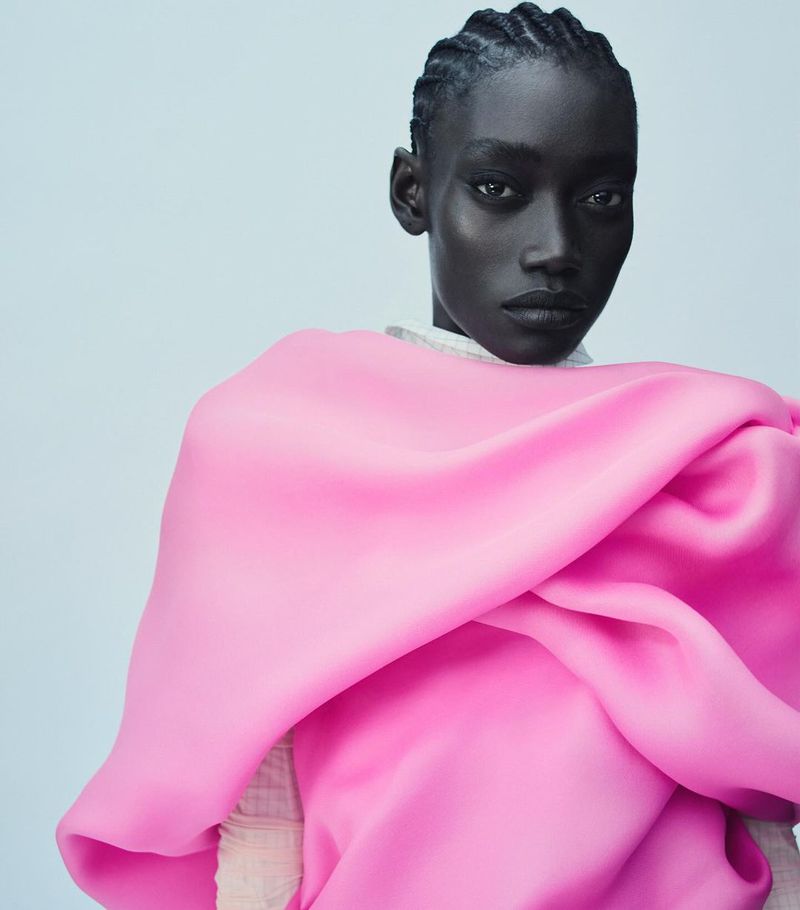Portrait of a Black woman in a pink fabric wrap against a light blue background, showcasing braids and neutral expression in a fashion setting.