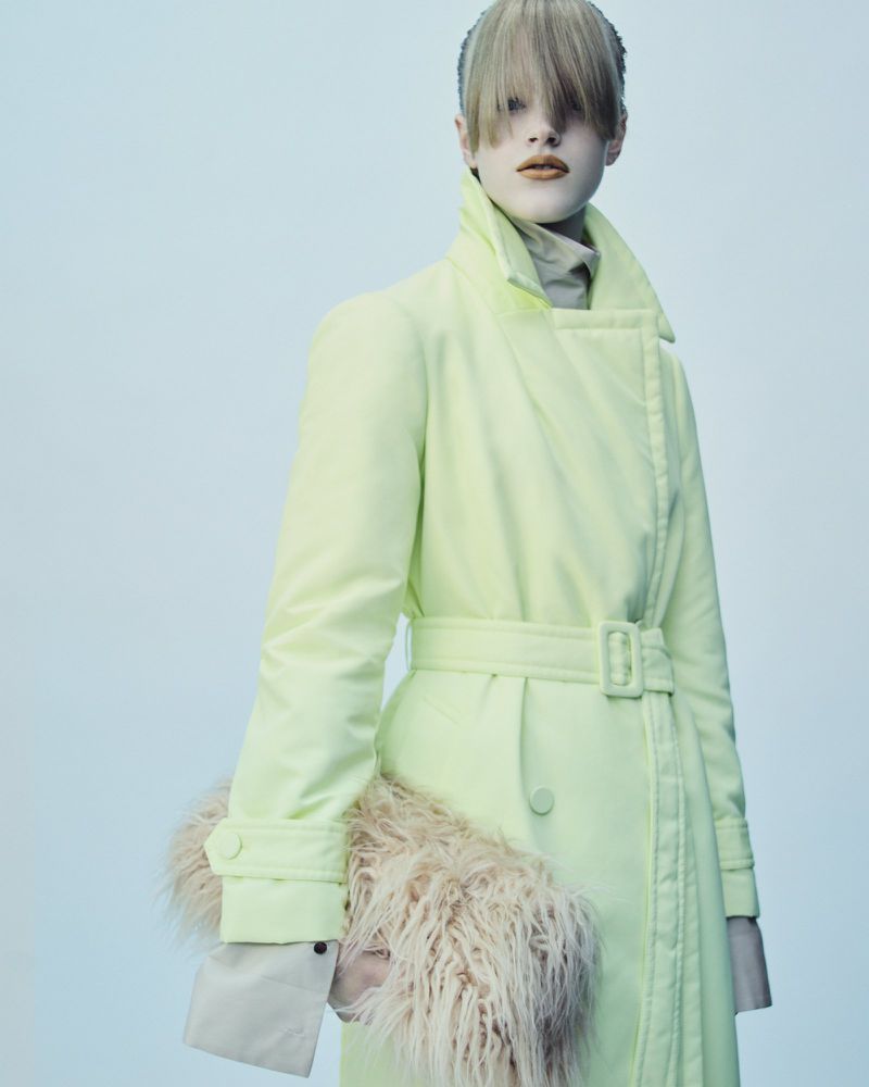 Fashion shot of a woman with blonde hair, wearing a light green trench coat and holding a beige fur accessory in a studio setting.