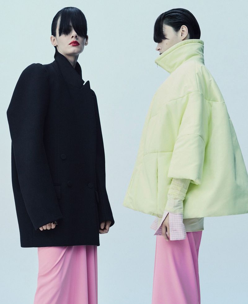 Two models posing in an oversized black coat and a pale yellow quilted jacket with pink skirts against a pale studio backdrop.