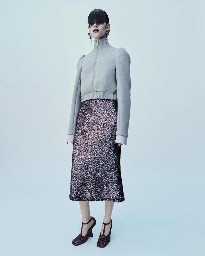 Full-length shot of a woman with dark hair wearing a gray jacket, multicolored sequin skirt, and t-strap heels against a light blue background.