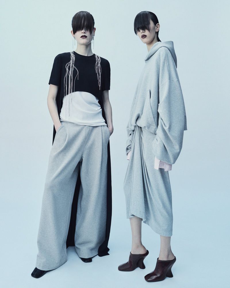Two young adult female fashion models pose in a studio wearing oversized grey and black clothing with long bangs covering their eyes.
