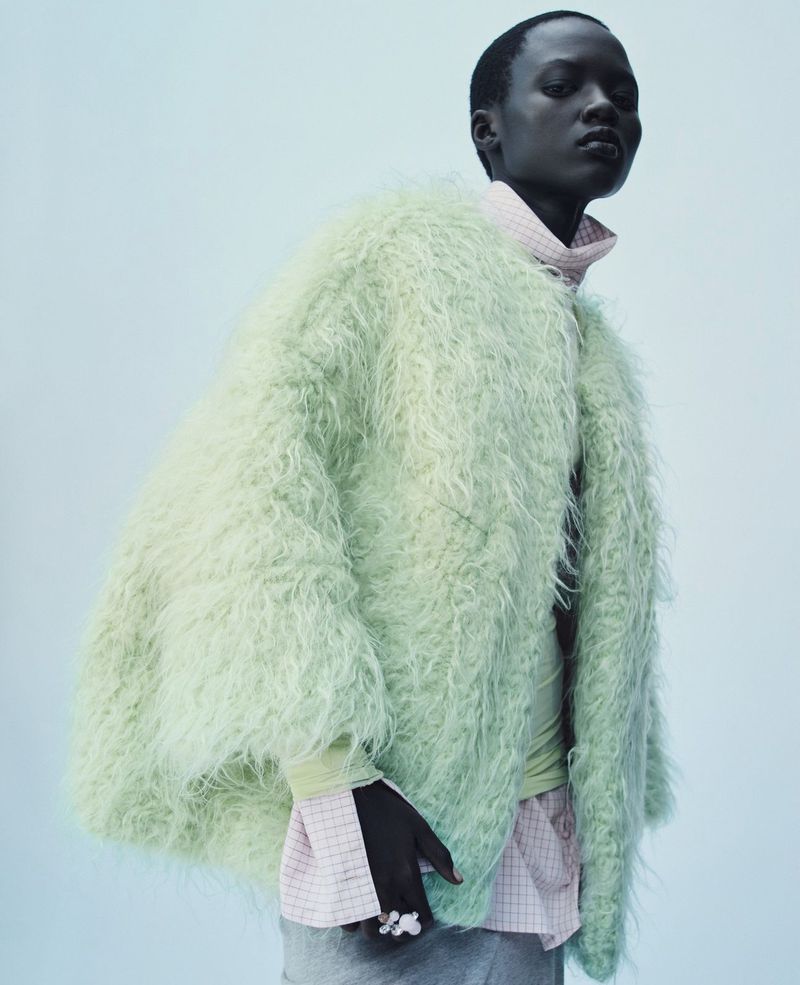 An adult female model with dark skin poses in a light-green faux fur coat, light-pink turtleneck, and grey skirt against a light-blue backdrop.