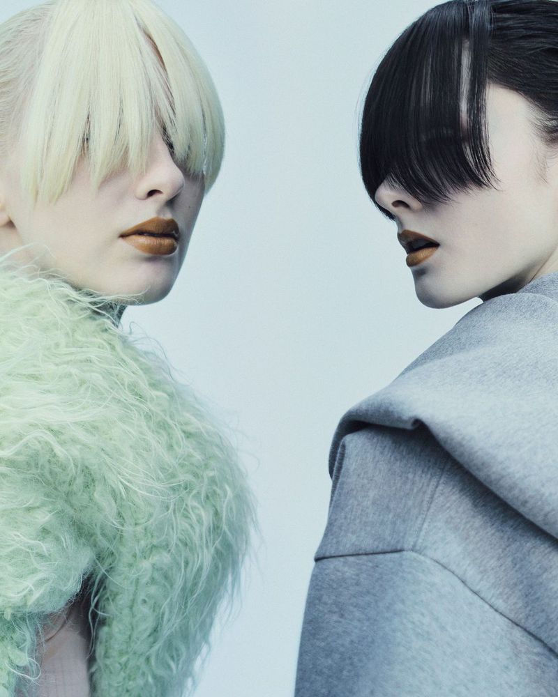 Two women with unique hairstyles and fashionable makeup pose in a close-up studio shot with soft lighting and a clean, light-blue background.