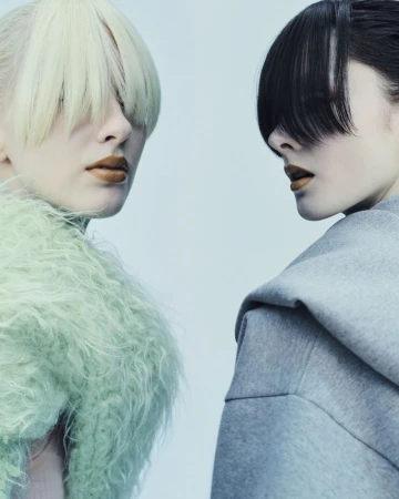 Two women with unique hairstyles and fashionable makeup pose in a close-up studio shot with soft lighting and a clean, light-blue background.