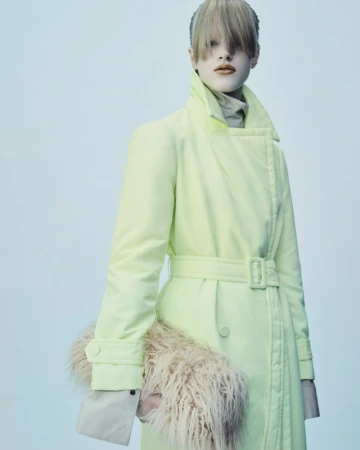 Model wearing a pale yellow belted coat and holding a fuzzy textured clutch against a light studio backdrop.