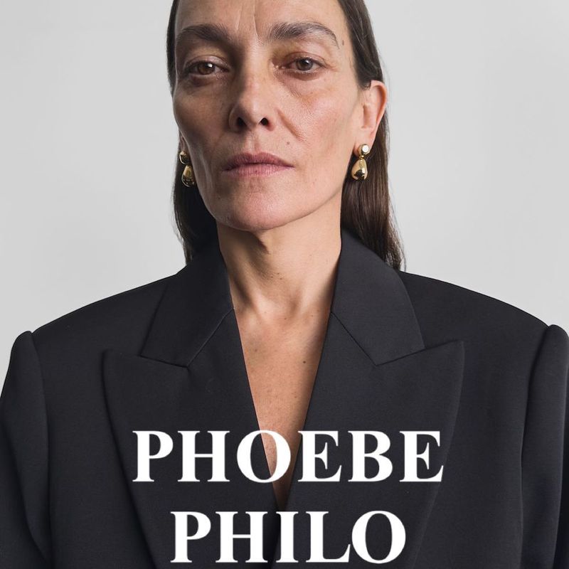 Adult woman in a black blazer and gold earrings poses in front of a white backdrop with "PHOEBE PHILO" text overlay.