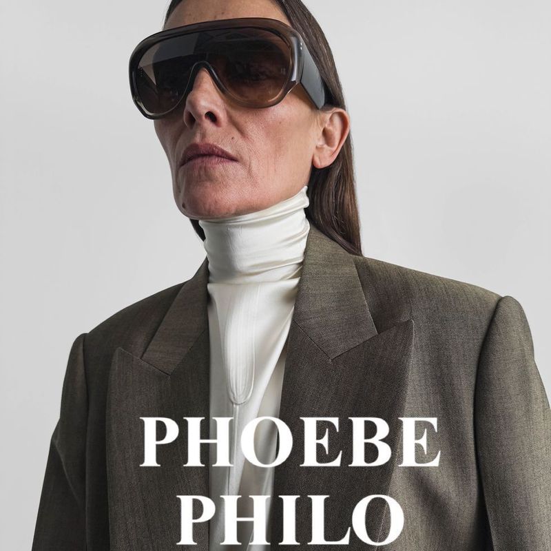 Fashion portrait of an adult woman wearing oversized sunglasses, a white turtleneck sweater, and a brown blazer against a neutral backdrop.