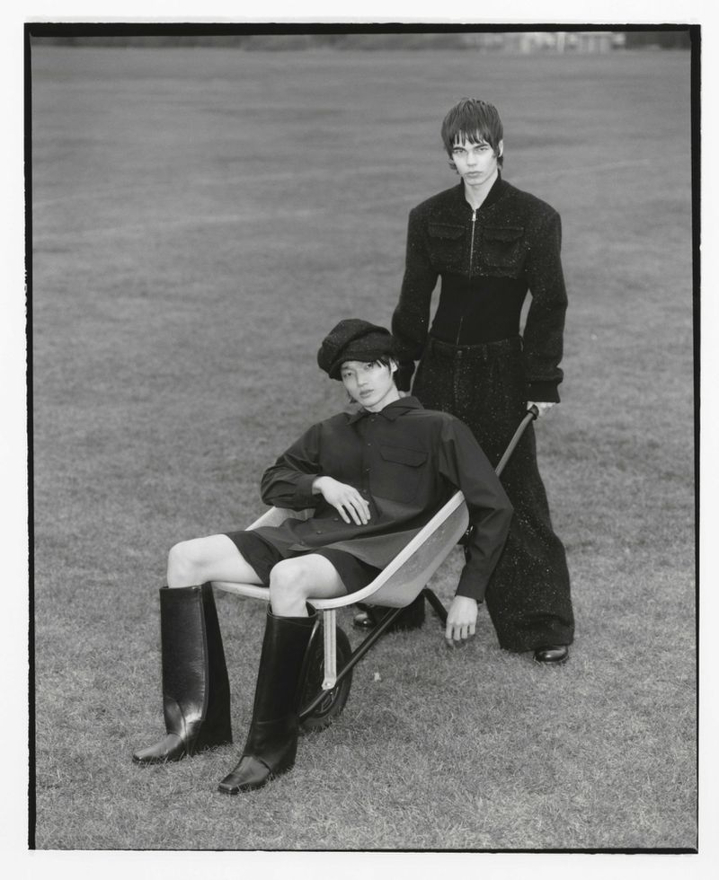 Monochrome fashion portrait of two adult women. One is seated in a wheelbarrow on a grass field, with the other woman standing behind.