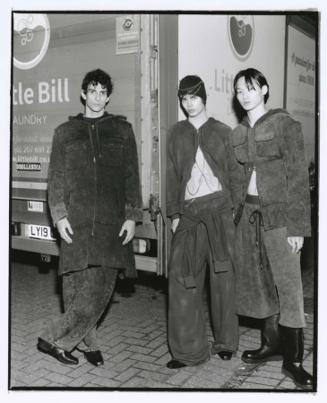 Three men pose outdoors in matching hooded jackets and trousers. The photo is in black and white with a commercial truck as the backdrop.