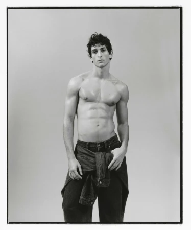 Striking black and white portrait of a muscular shirtless man in denim pants and jacket, standing in a photo studio.