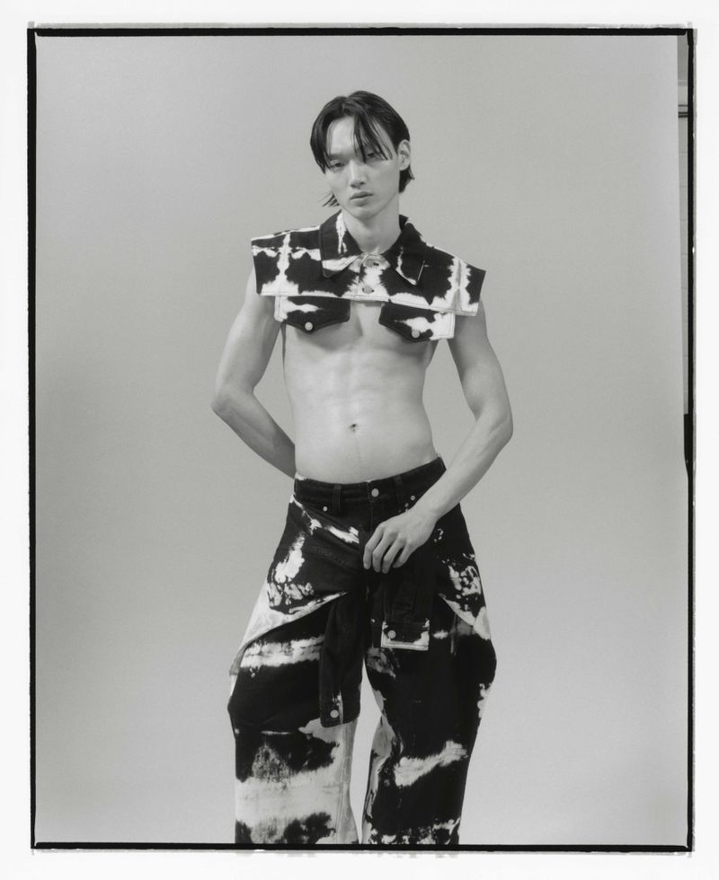 Monochrome studio shot of a slender adult male model wearing patterned crop top and pants, posing against a neutral backdrop.
