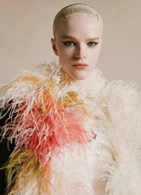 Close-up portrait of a young adult woman with pale skin, wearing a tan wig cap and a feathered shawl with pink and yellow accents in studio lighting.