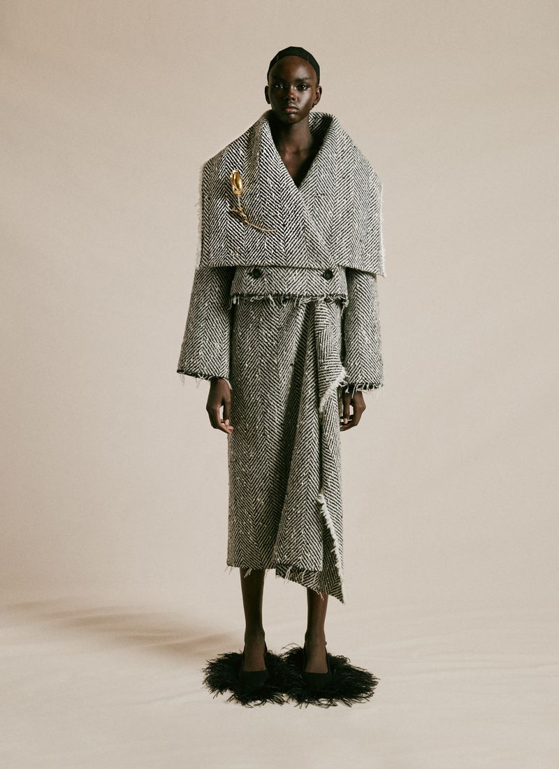 A full-length portrait of a Black female fashion model wearing an oversized grey and white tweed suit with an A-line skirt and feather shoes.