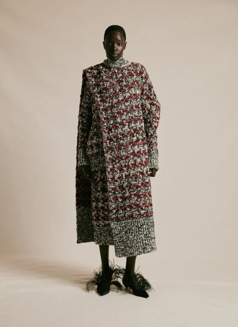 Full-length portrait of a Black woman in a textured long-sleeved dress with feathered heels, against a neutral studio backdrop.
