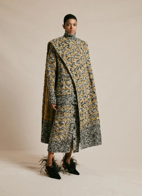 Full-length portrait of a Black fashion model in a textured yellow and black coat, skirt, and feather-trimmed boots on a neutral backdrop.