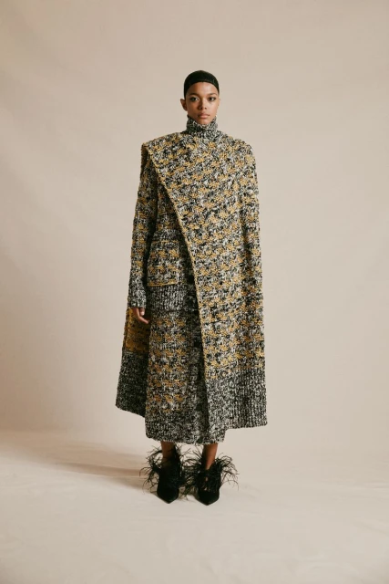 A fashionable adult female poses in a tweed coat and skirt with feathered heels against a neutral backdrop in a studio setting.