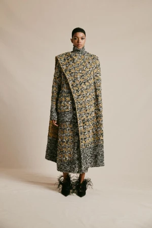 A fashionable adult female poses in a tweed coat and skirt with feathered heels against a neutral backdrop in a studio setting.