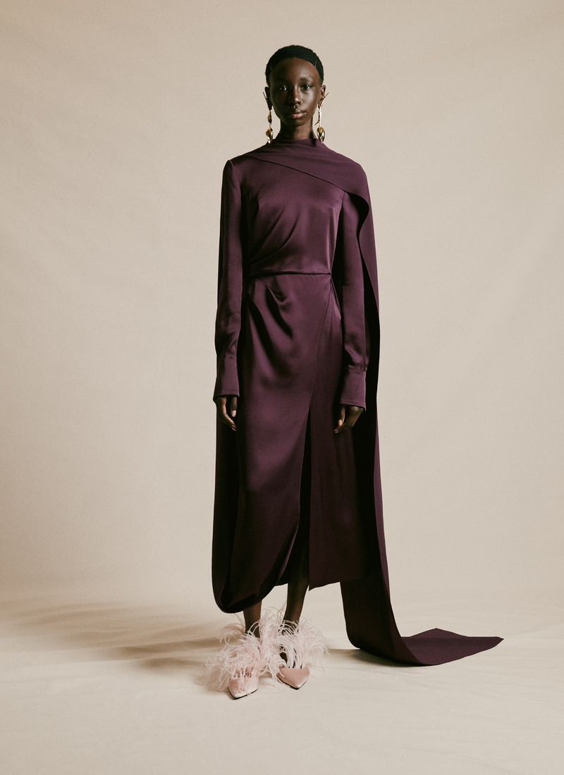 A single woman poses in a photo studio, wearing a plum-colored satin dress and fuzzy heels, illuminated by bright, even lighting.