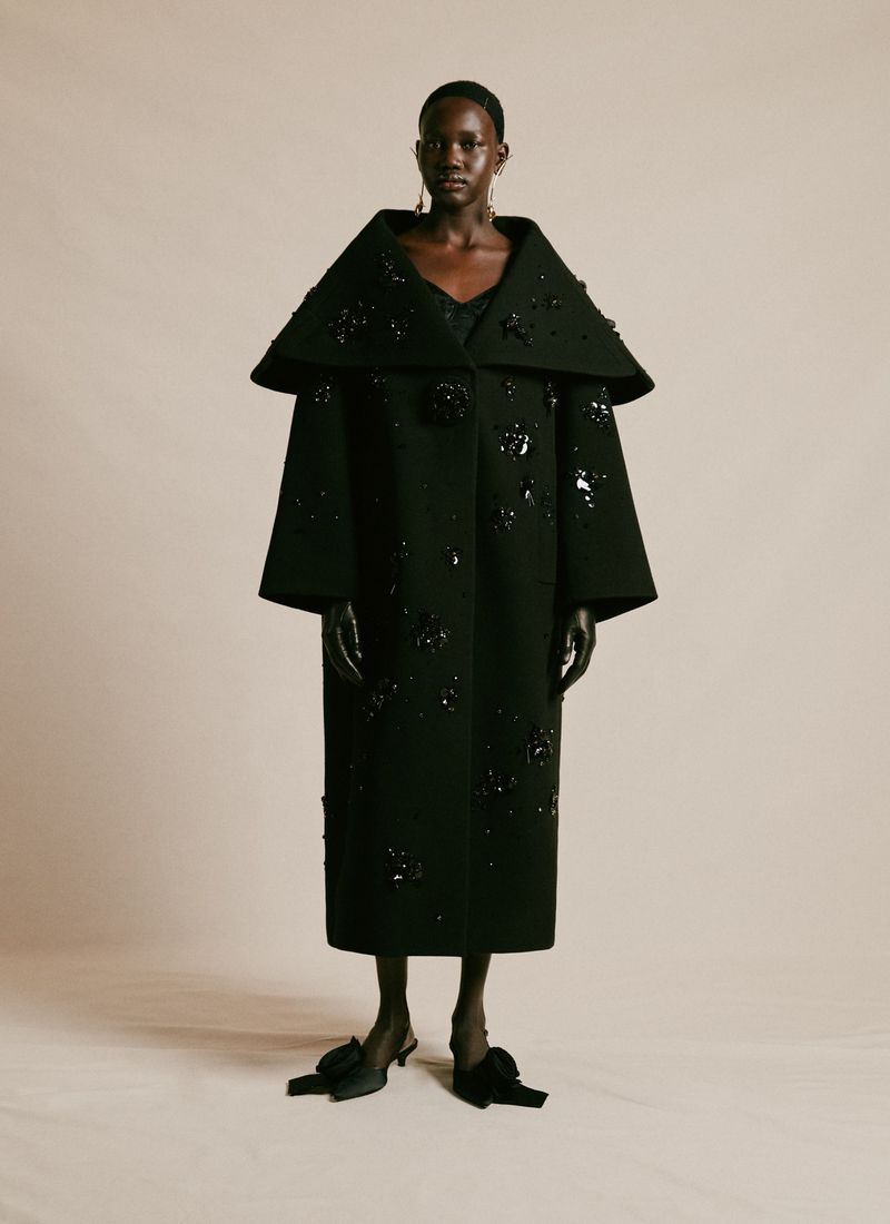 An African-American woman models a long black wool coat adorned with black flower appliqués, black gloves and black bow shoes against a studio backdrop.