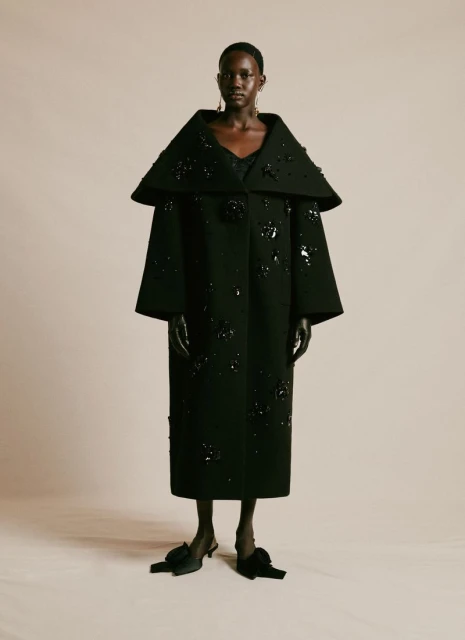 An African-American woman models a long black wool coat adorned with black flower appliqués, black gloves and black bow shoes against a studio backdrop.