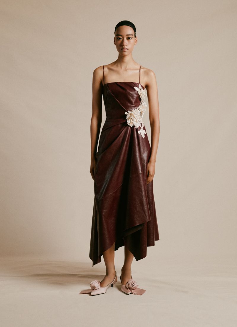 Woman in a burgundy faux leather dress with thin straps and floral embellishments poses in a studio with neutral lighting.