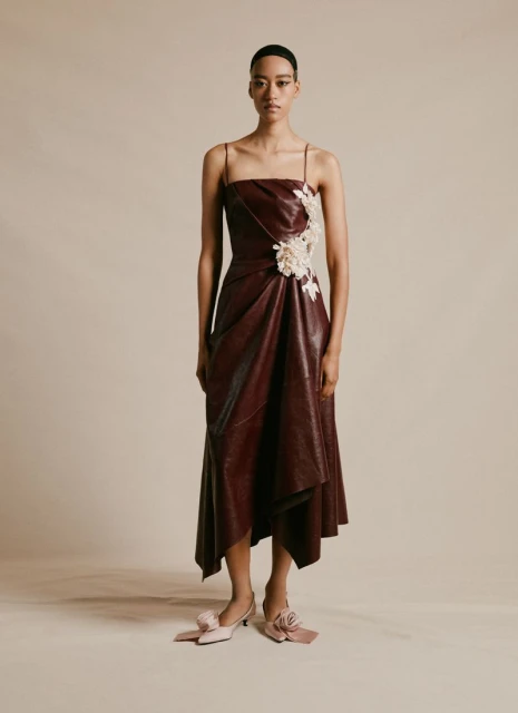 Woman in a burgundy faux leather dress with thin straps and floral embellishments poses in a studio with neutral lighting.