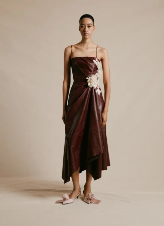Woman in a burgundy faux leather dress with thin straps and floral embellishments poses in a studio with neutral lighting.