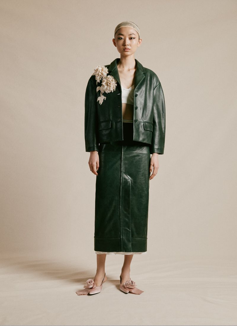 An Asian woman models a dark green leather jacket and skirt with floral accents, accessorized with matching shoes, posing in studio.