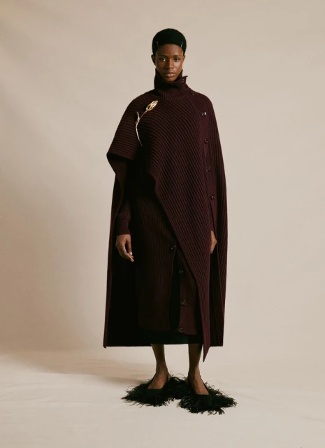 A full-length portrait captures a Black woman in a burgundy knit dress and cape with feather slippers against a neutral backdrop.