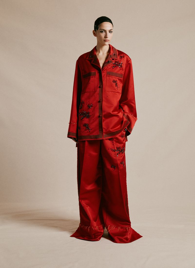 Fashion model stands in a photo studio wearing a red satin pajama set decorated with floral embellishments, against a neutral backdrop.