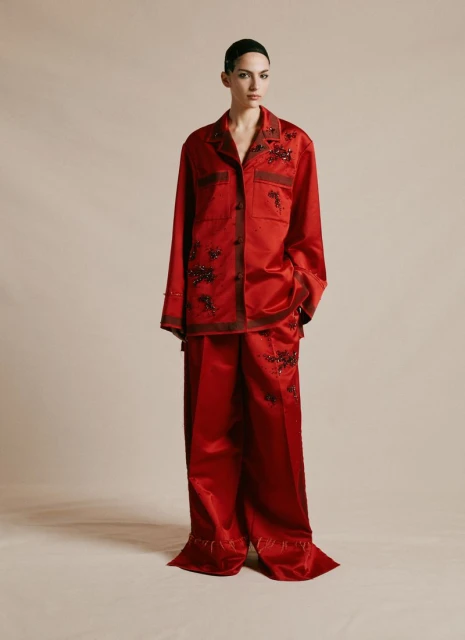 Fashion model stands in a photo studio wearing a red satin pajama set decorated with floral embellishments, against a neutral backdrop.